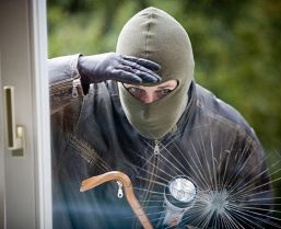 Ein Einbrecher mit Maske und Handschuhen schaut durch ein zerbrochenes Fenster